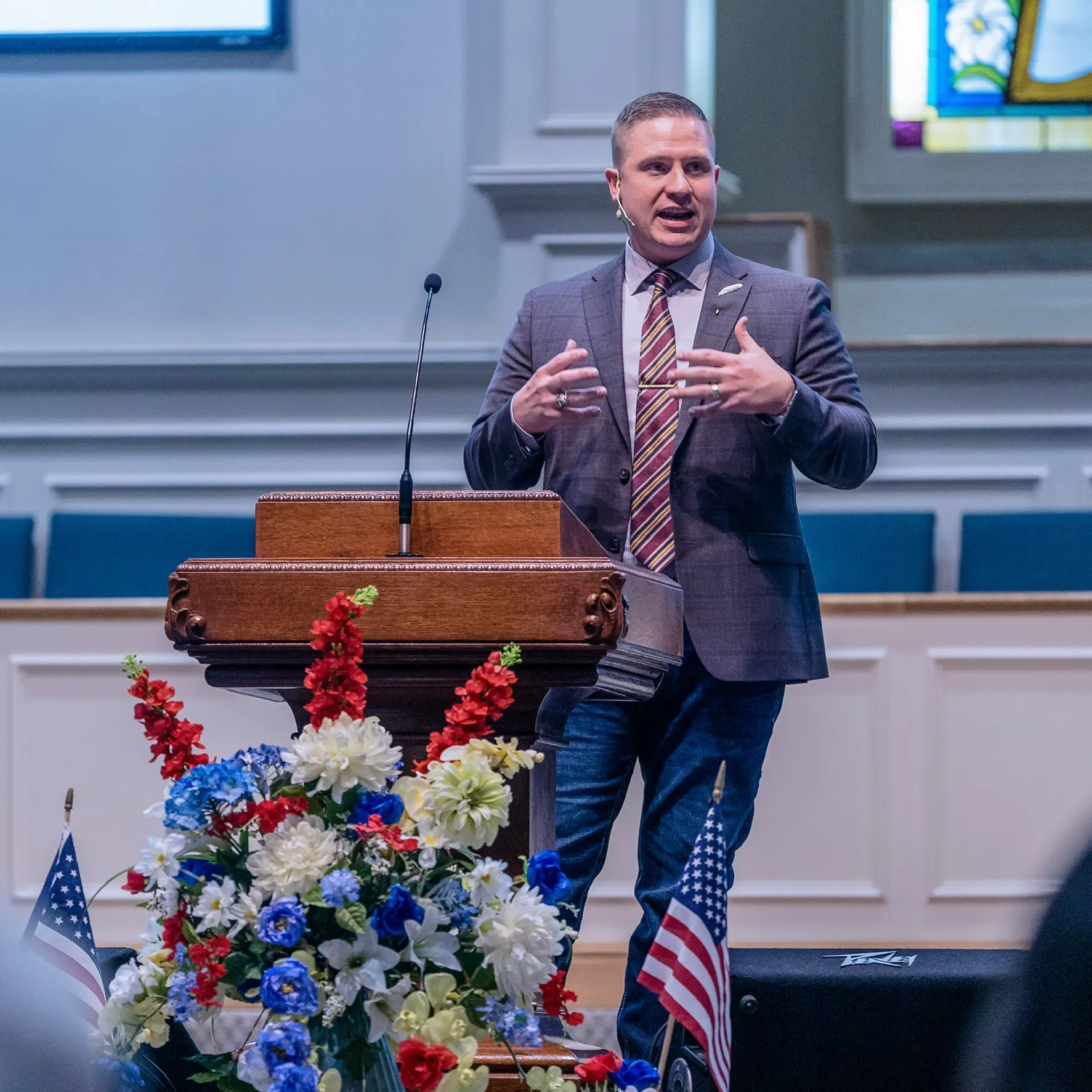 Aaron speaking beside a wooden pulpit with flowers and American flags